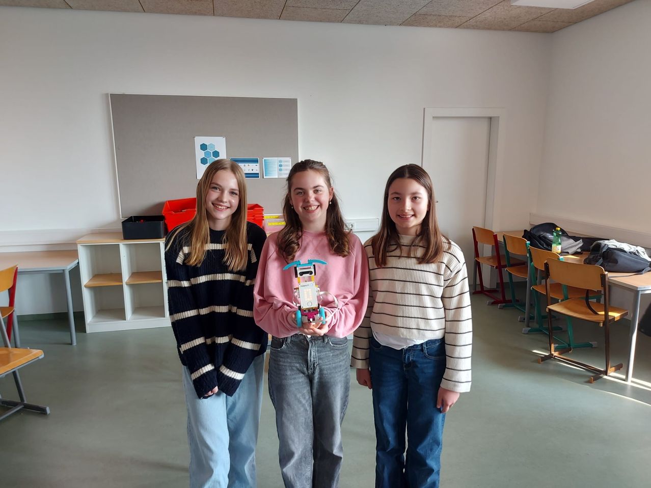 Drei junge Mädchen stehen in einem Raum mit einer grauen Wand und einem weißen Regal. Das Mädchen in der Mitte hält ein Spielzeug. Sie lächeln und posieren für ein Foto.