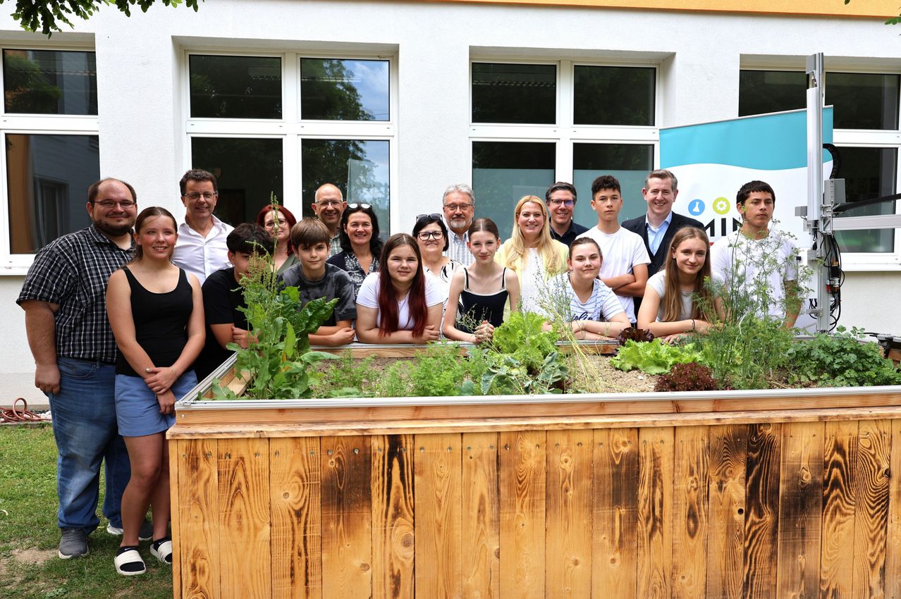 Eine Gruppe von Menschen posiert vor einem hölzernen Pflanzkasten mit Grünpflanzen vor einem weißen Gebäude.