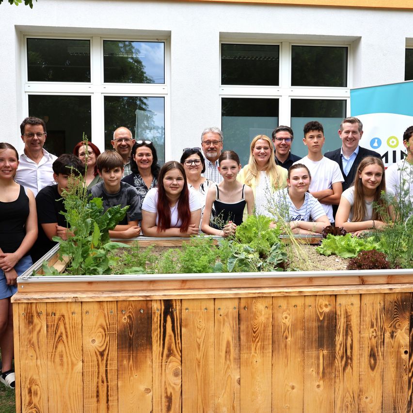 Eine Gruppe von Menschen posiert vor einem hölzernen Pflanzkasten mit Grünpflanzen vor einem weißen Gebäude.