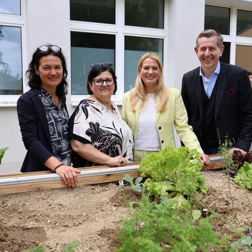Vier Erwachsene stehen hinter einem Gartenbeet und lächeln in die Kamera. Sie stehen vor einem weißen Gebäude mit großen Fenstern.