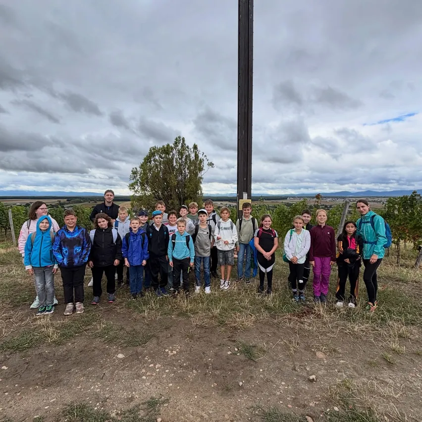 Eine Gruppe von Kindern und Erwachsenen posiert für ein Foto vor einem großen Kreuz auf einem Hügel unter einem bewölkten Himmel.