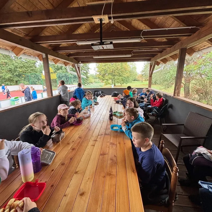 Eine Gruppe von Kindern sitzt an einem langen Holztisch in einem Pavillon und isst und trinkt.
