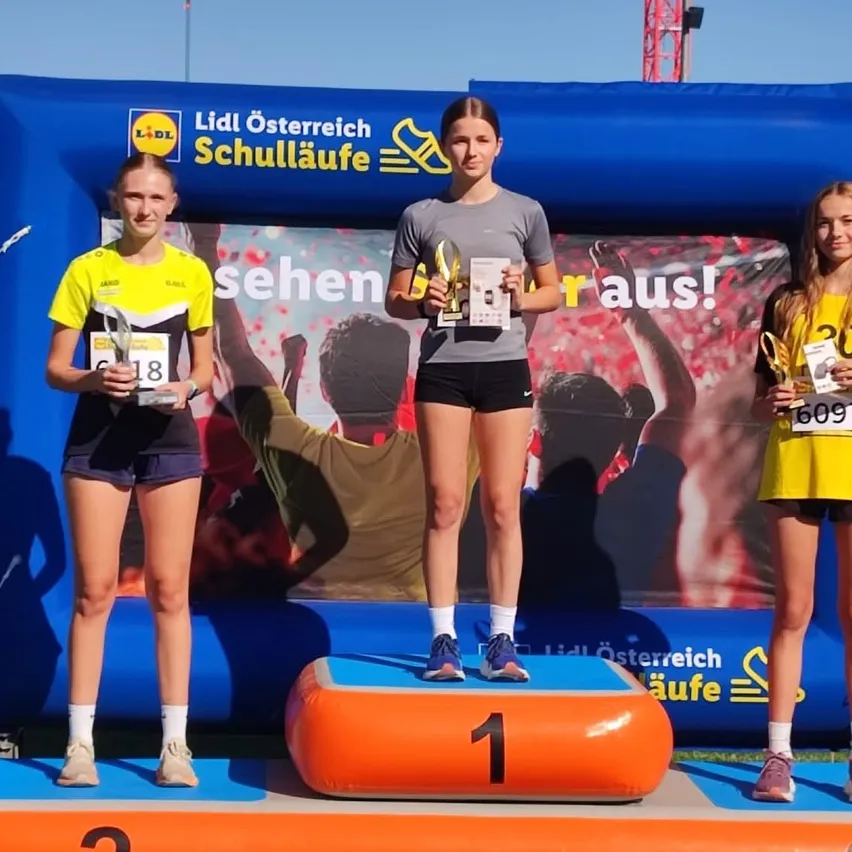 Drei Frauen stehen auf einem Podest und halten Trophäen. Die Frau in der Mitte trägt ein graues T-Shirt und schwarze Shorts. Die Frau auf der linken Seite trägt ein gelbes T-Shirt und schwarze Shorts. Die Frau auf der rechten Seite trägt ein gelbes T-Shirt und schwarze Shorts. Hinter ihnen steht ein blaues Aufblasbares mit einem großen Plakat.
