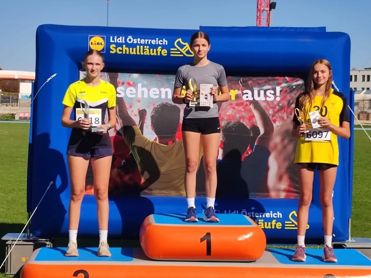 Drei Frauen stehen auf einem Podest und halten Trophäen. Die Frau in der Mitte trägt ein graues T-Shirt und schwarze Shorts. Die Frau auf der linken Seite trägt ein gelbes T-Shirt und schwarze Shorts. Die Frau auf der rechten Seite trägt ein gelbes T-Shirt und schwarze Shorts. Hinter ihnen steht ein blaues Aufblasbares mit einem großen Plakat.