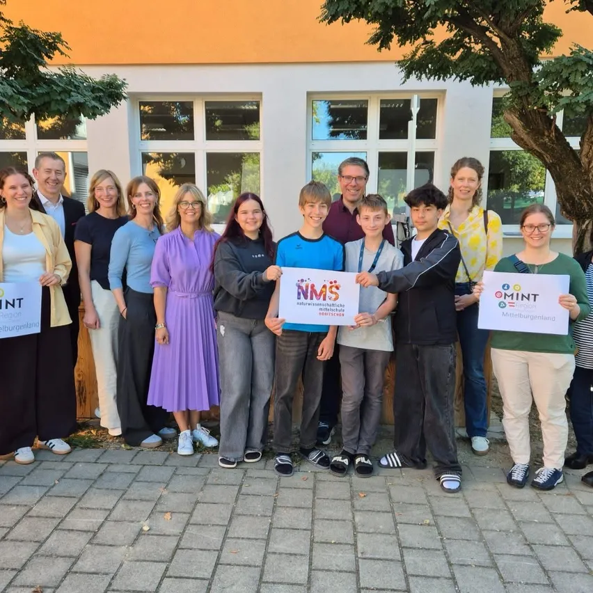Eine Gruppe von Menschen steht vor einem Gebäude und hält Schilder mit dem Text 'MINT' und 'NMS'.
