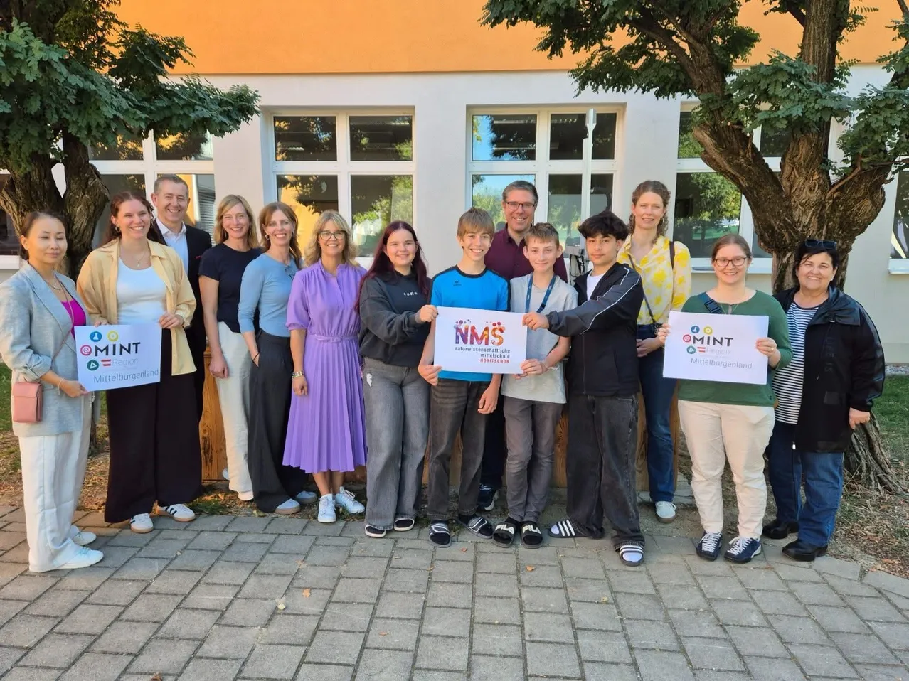 Eine Gruppe von Menschen steht vor einem Gebäude und hält Schilder mit dem Text 'MINT' und 'NMS'.