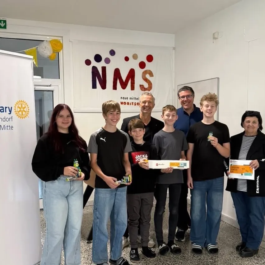 Eine Gruppe von Menschen steht zusammen in einem Raum, lächelt und posiert für ein Foto. Sie halten Schachteln und Spielzeug. Hinter ihnen ist ein Banner mit dem Text 'NMS' und einem Logo.