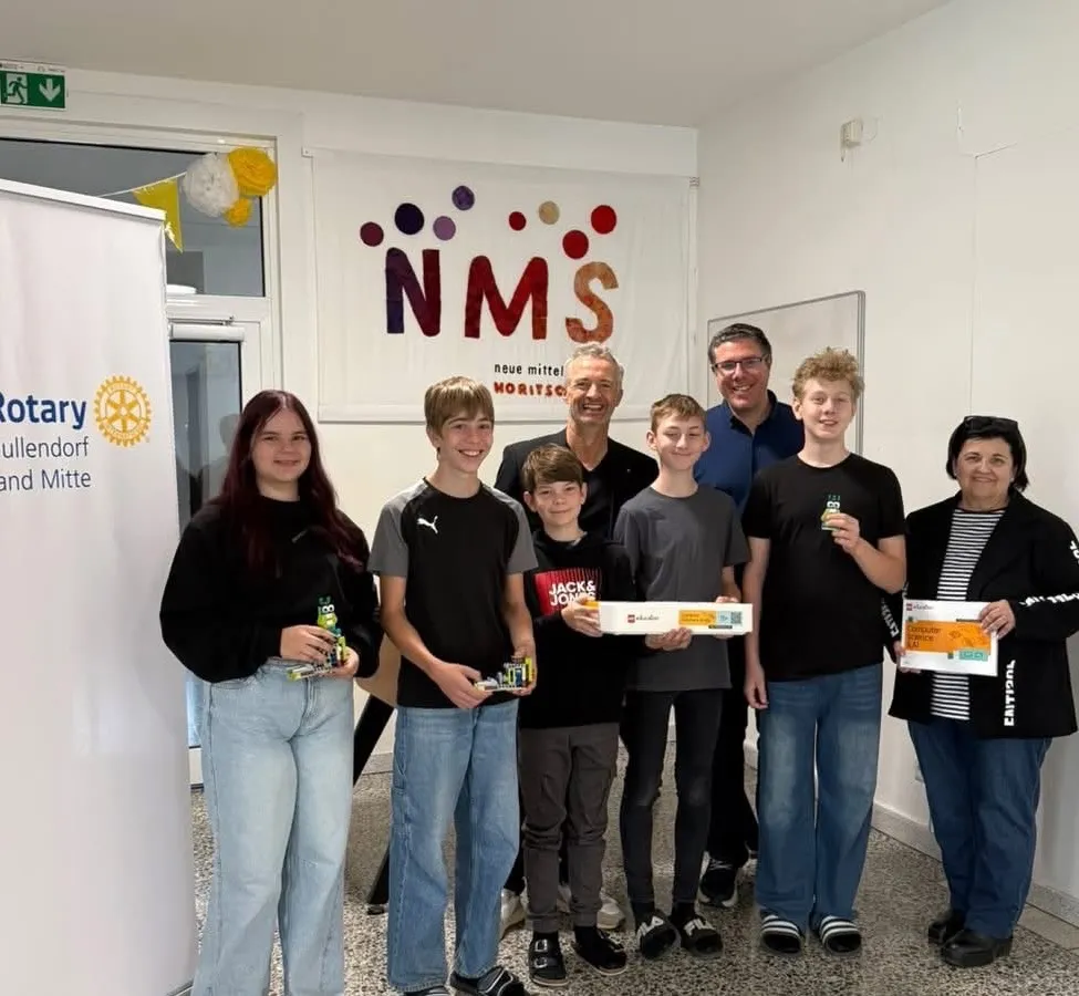 Eine Gruppe von Menschen steht zusammen in einem Raum, lächelt und posiert für ein Foto. Sie halten Schachteln und Spielzeug. Hinter ihnen ist ein Banner mit dem Text 'NMS' und einem Logo.