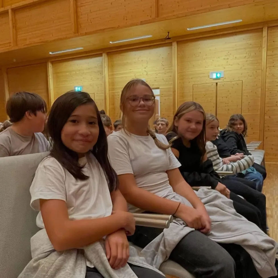 Eine Gruppe junger Mädchen und Jungen sitzt in Stühlen in einem Raum mit Holzwänden.
