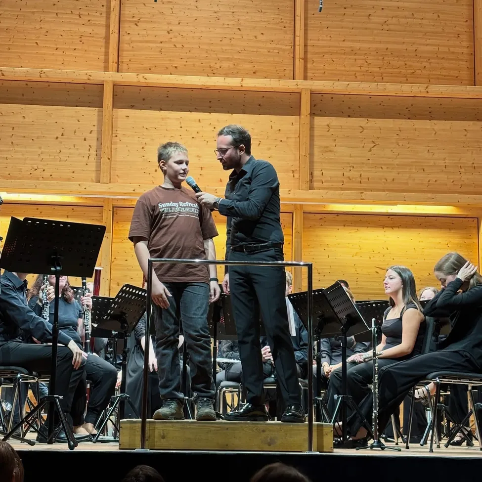 Ein Mann steht auf der Bühne mit einem Jungen, und sie halten beide ein Mikrofon. Der Mann trägt eine Brille und einen Anzug, während der Junge ein braunes Hemd trägt. Hinter ihnen ist eine Holzwand mit einer Reihe von Musikinstrumenten auf dem Boden.