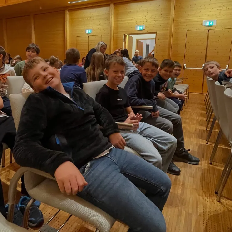 Eine Gruppe von Kindern sitzt in Stühlen in einem Raum. Der Junge vorne lächelt und schaut in die Kamera.