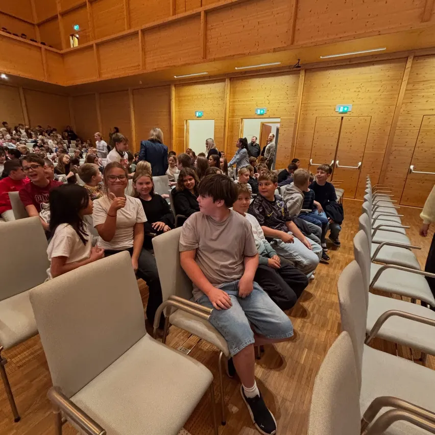 Eine Gruppe von Kindern und Erwachsenen sitzt in Stühlen in einem großen Raum. Der Raum hat Holzwände und -böden und es gibt Türen auf der rechten Seite.