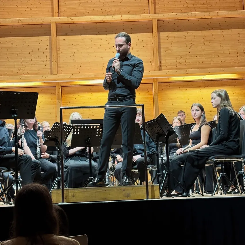 Ein Mann steht auf der Bühne mit einem Mikrofon, möglicherweise hält er eine Rede oder singt. Hinter ihm spielen eine Gruppe von Musikern ihre Instrumente.