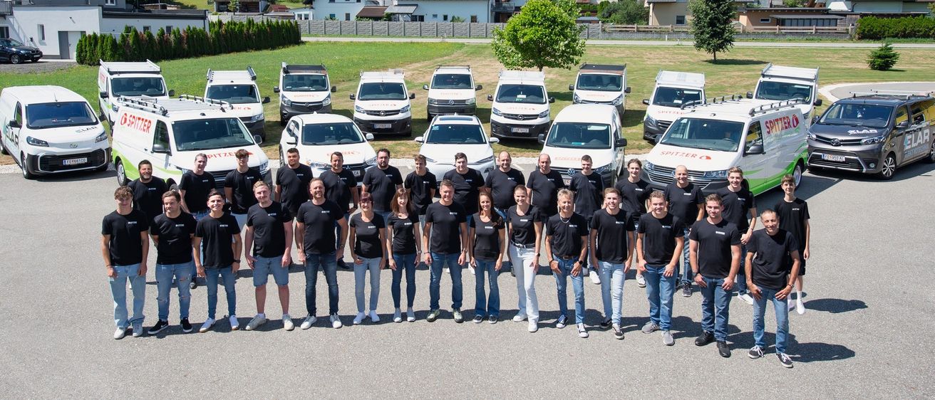 Eine Gruppe von Menschen in passenden schwarzen T-Shirts posiert vor einer Reihe weißer Lieferwagen mit dem Wort 'Spitzen' an den Seiten. Dahinter sind Häuser in der Ferne zu sehen.