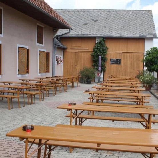 Ein rustikaler Außenbereich mit Holzbänken und Tischen in Reihen, neben einem Gebäude mit Holztüren und Fenstern, umgeben von Pflanzen und einem Ziegelboden.