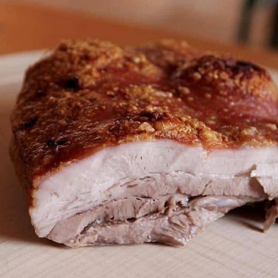 Ein Nahaufnahme eines geschnittenen Stücks gebratenen Schweinebauchs mit einer knusprigen, goldbraunen Kruste und einem saftigen, rosafarbenen Inneren. Das Fleisch ist geschnitten, um seine Schichten zu zeigen und den Kontrast zwischen der knusprigen Außenseite und dem zarten, geschmackvollen Inneren zu demonstrieren.