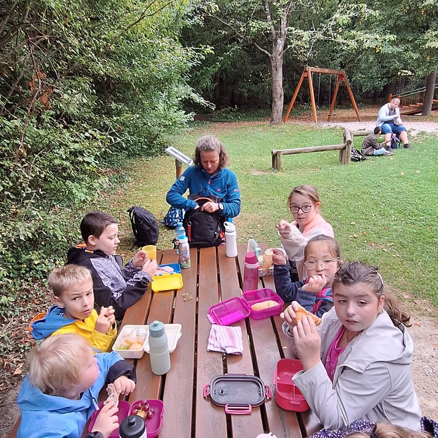 Eine Gruppe von Kindern und eine erwachsene Frau sitzen an einem Picknicktisch und essen. Der Tisch ist mit Flaschen, Lunchboxen und Servietten gedeckt. Hinter ihnen ist ein Spielplatz mit Schaukel und Rutsche zu sehen.