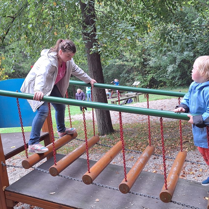 Zwei Kinder klettern eine Holzleiter in einem Park. Ein Kind schaut in die Kamera. Sie sind von Bäumen umgeben.