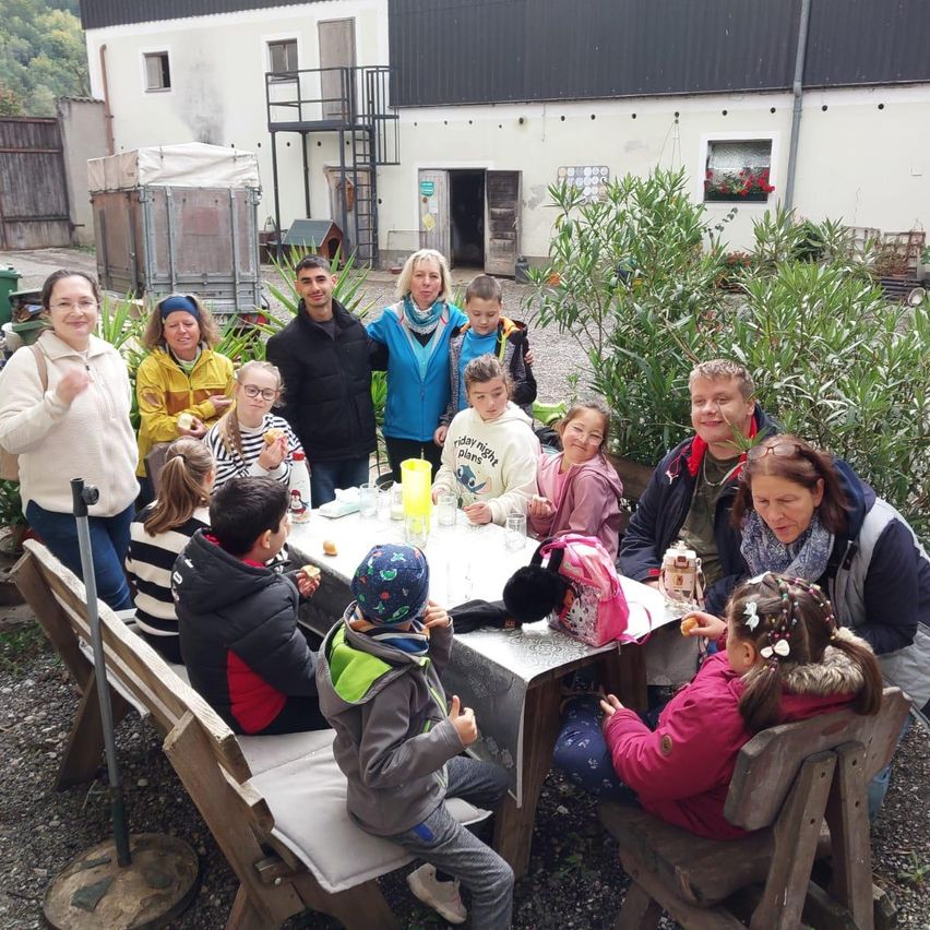 Eine Gruppe von Menschen sitzt draußen am Tisch, einige mit Essen in den Händen. Dahinter ist ein Gebäude und Grünpflanzen zu sehen.