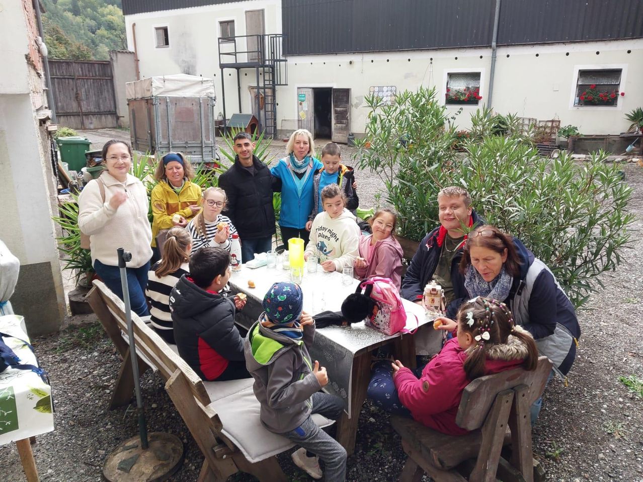 Eine Gruppe von Menschen sitzt draußen am Tisch, einige mit Essen in den Händen. Dahinter ist ein Gebäude und Grünpflanzen zu sehen.