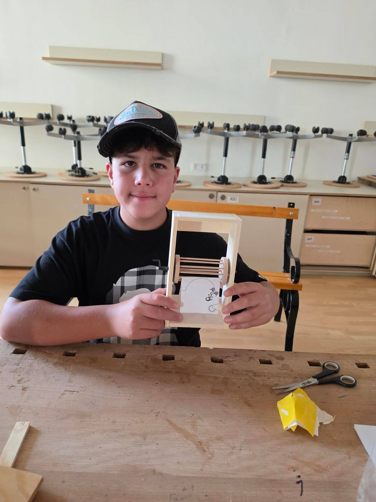 Ein junger Junge hält ein kleines hölzerne Webstuhl und lächelt in einer Werkstatt mit Holzbänken und Schränken im Hintergrund.