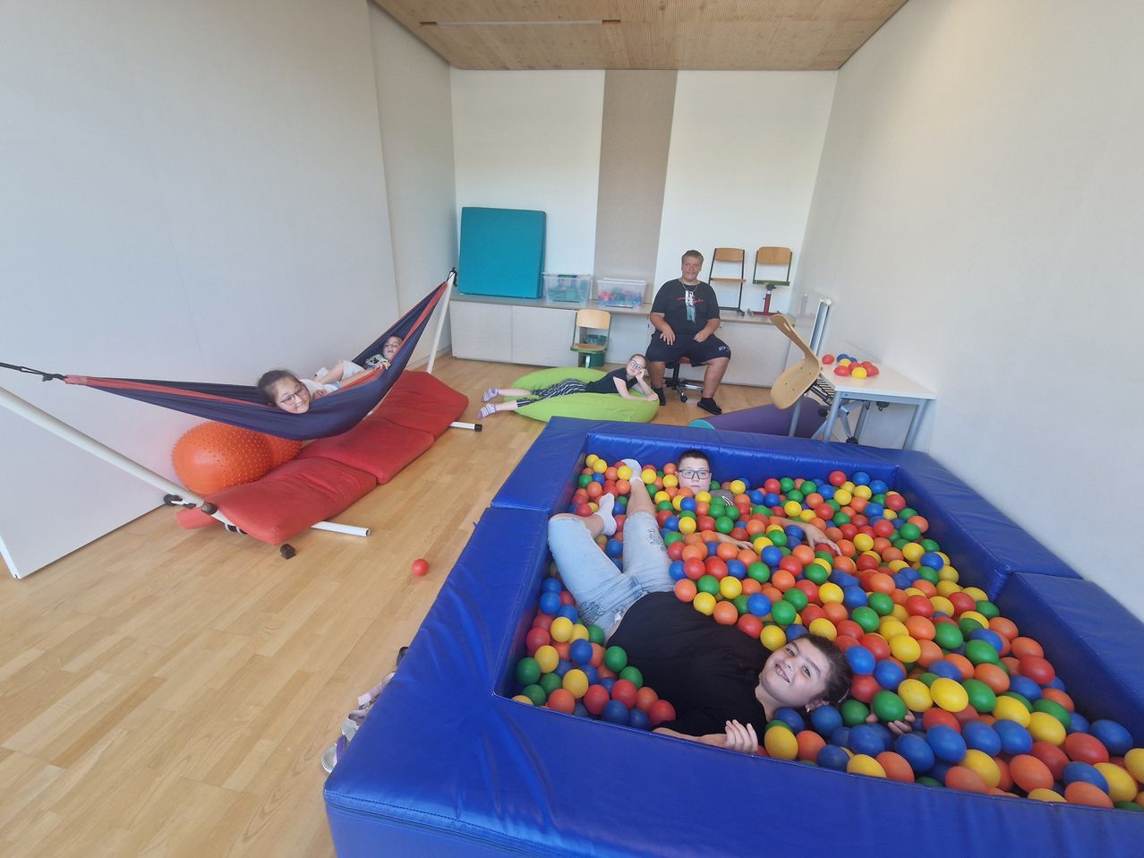 Vier Kinder spielen in einer Ballgrube, eines liegt herum, während ein anderes in einer Hängematte ist. Ein Mann sitzt in der Nähe.