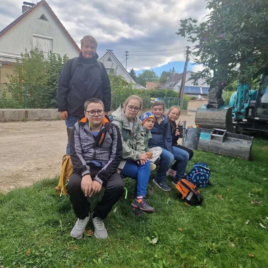Eine Gruppe von Kindern und zwei Erwachsenen sitzen auf dem Gras. Sie tragen Jacken und einige tragen Brillen. Es gibt Rucksäcke und einen Becher auf dem Gras. Hinter ihnen steht ein Haus mit einem Zaun und einem Baufahrzeug.