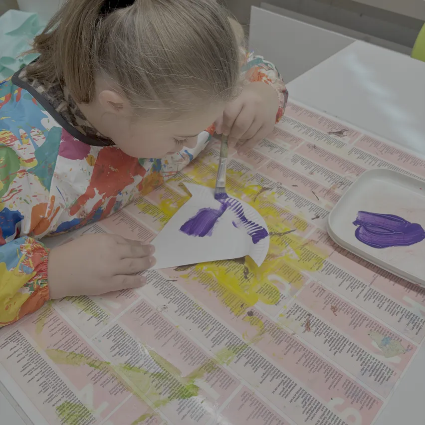 Ein junges Mädchen malt ein weißes Papier mit lila Farbe auf einem Tisch mit einer Zeitung darunter.