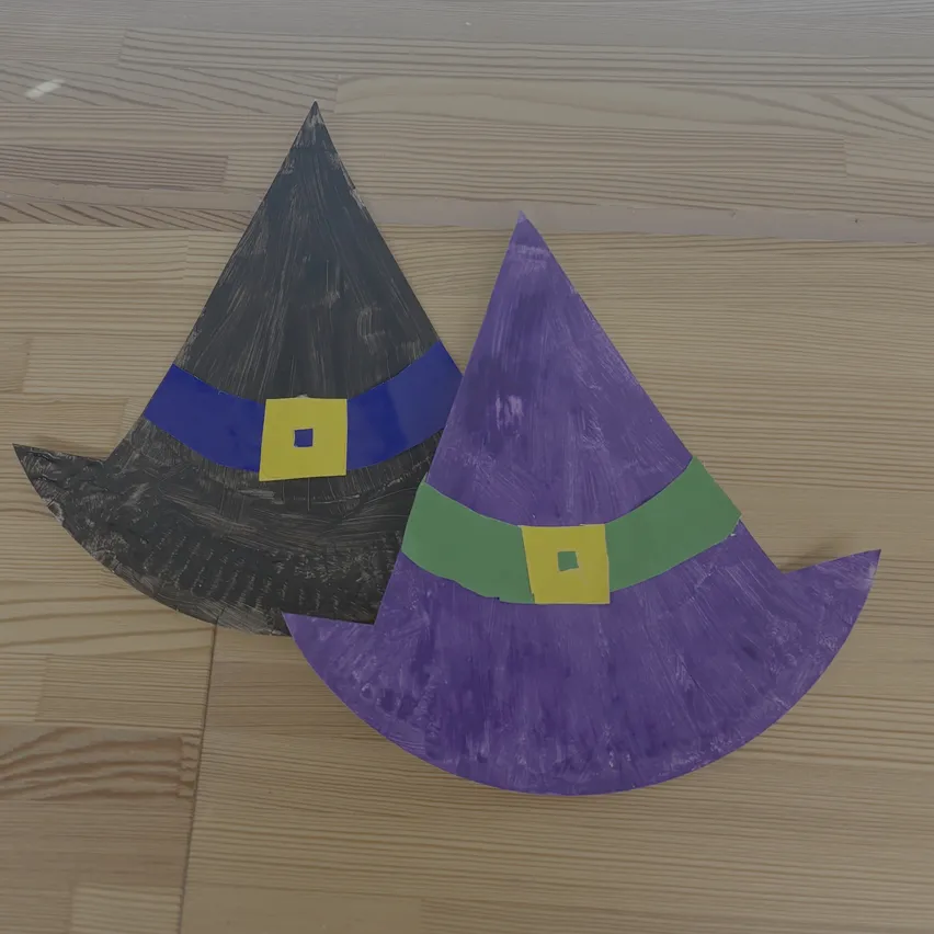 Zwei Papierhexenhüte auf einer Holzoberfläche. Einer ist lila mit einem grünen Band und einem gelben Quadrat, der andere ist schwarz mit einem blauen Band und einem gelben Quadrat.