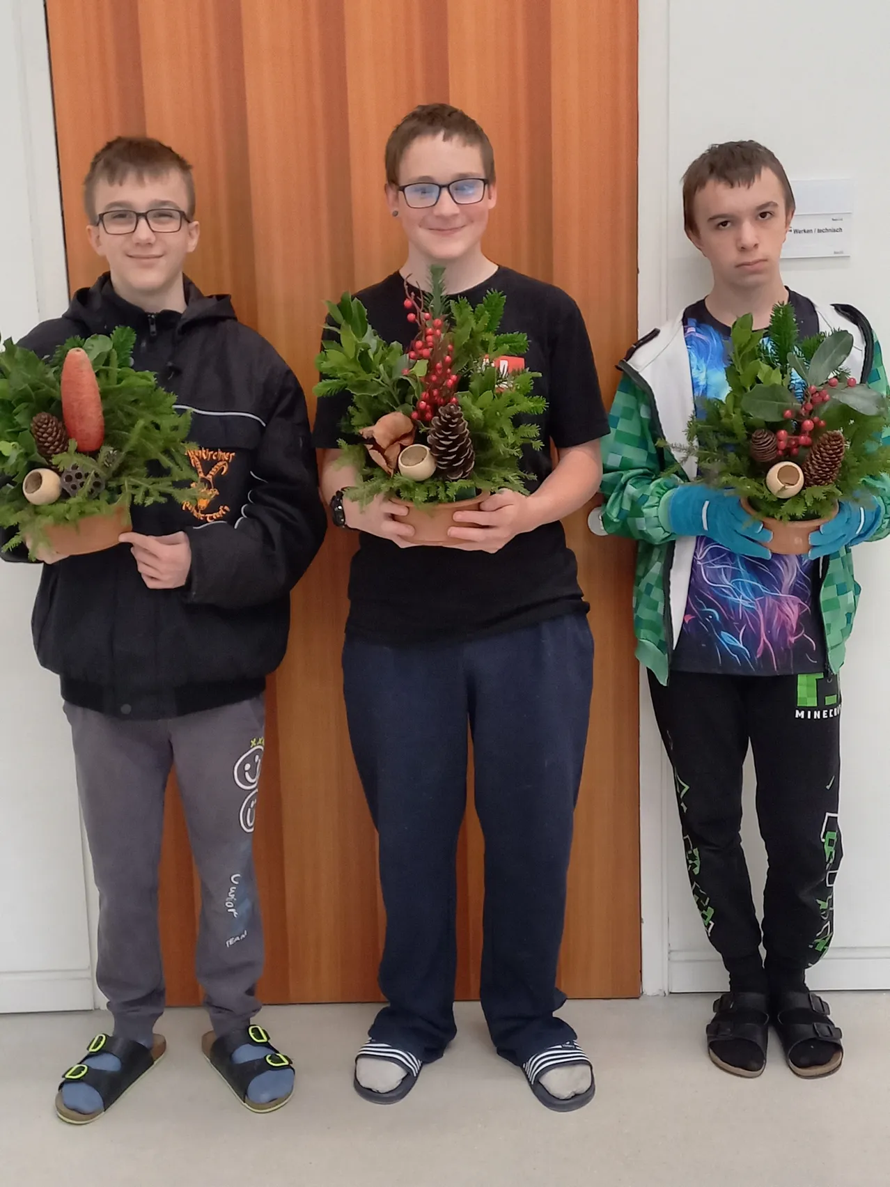 Drei junge Jungen stehen zusammen und halten jeweils eine dekorative Girlande aus Grün und Tannenzapfen. Sie stehen vor einer Holztür.