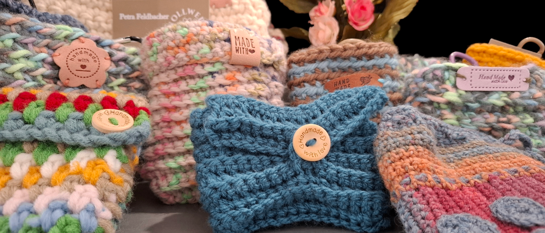 Eine Sammlung von farbenfrohen gestrickten Artikeln ist auf einer grauen Oberfläche ausgestellt. Ein Strauß rosa Blumen steht hinter den Artikeln. Jeder Artikel hat ein einzigartiges Design und ein selbstgemachtes Etikett.