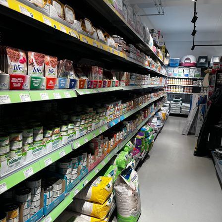 Ein Supermarktgang mit verschiedenen Produkten in den Regalen, einschließlich Konserven, Tierfutter und andere Artikel. Die Regale sind gut bestückt mit einer Vielzahl von Produkten.
