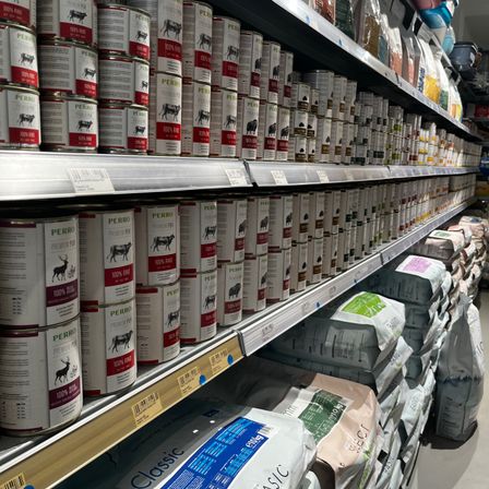 Ein Supermarktregal mit Reihen von Dosenfutter für Haustiere. Jede Dose hat ein Etikett mit einem Bild eines Tieres und Text, der den Produktnamen und den Fleischanteil angibt. Unter den Dosen befinden sich Säcke mit Tierfutter.