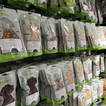 Eine Ausstellung von Hundefuttertüten hängt in einem Ladenregal. Die Tüten sind mit verschiedenen Hundefutternamen und -arten beschriftet. Die Tüten sind grün und weiß mit Hundeillustrationen. Die Tüten sind in verschiedenen Größen und Gewichten.