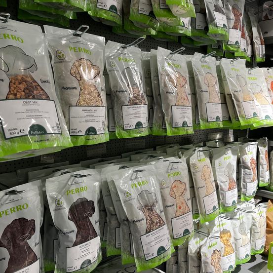 Eine Ausstellung von Hundefuttertüten hängt in einem Ladenregal. Die Tüten sind mit verschiedenen Hundefutternamen und -arten beschriftet. Die Tüten sind grün und weiß mit Hundeillustrationen. Die Tüten sind in verschiedenen Größen und Gewichten.
