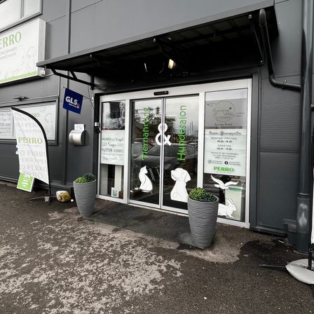 Ein graues Gebäude mit einem Vordach und Glastüren, beschriftet mit 'Remanah & Hundesalon'. Vor dem Eingang stehen zwei Topfpflanzen. Auf der linken Seite sind Banner angebracht.