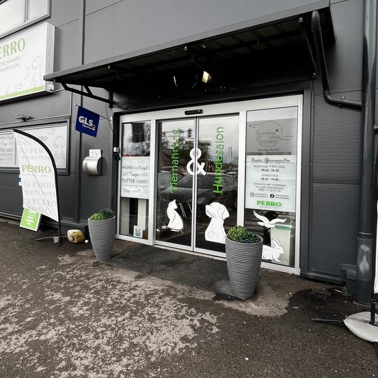 Ein graues Gebäude mit einem Vordach und Glastüren, beschriftet mit 'Remanah & Hundesalon'. Vor dem Eingang stehen zwei Topfpflanzen. Auf der linken Seite sind Banner angebracht.