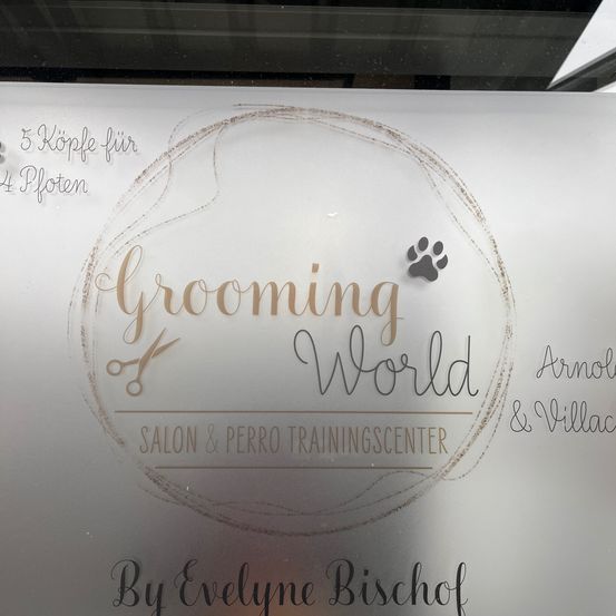 Ein weißer Schild zeigt ein Logo für Grooming World, bestehend aus einem Kreis mit einem Pfotenabdruck und einer Schere. Der Schild trägt den Text 'Grooming World Salon & Perro Trainingscenter' und ist von Evelyne Bischof signiert.