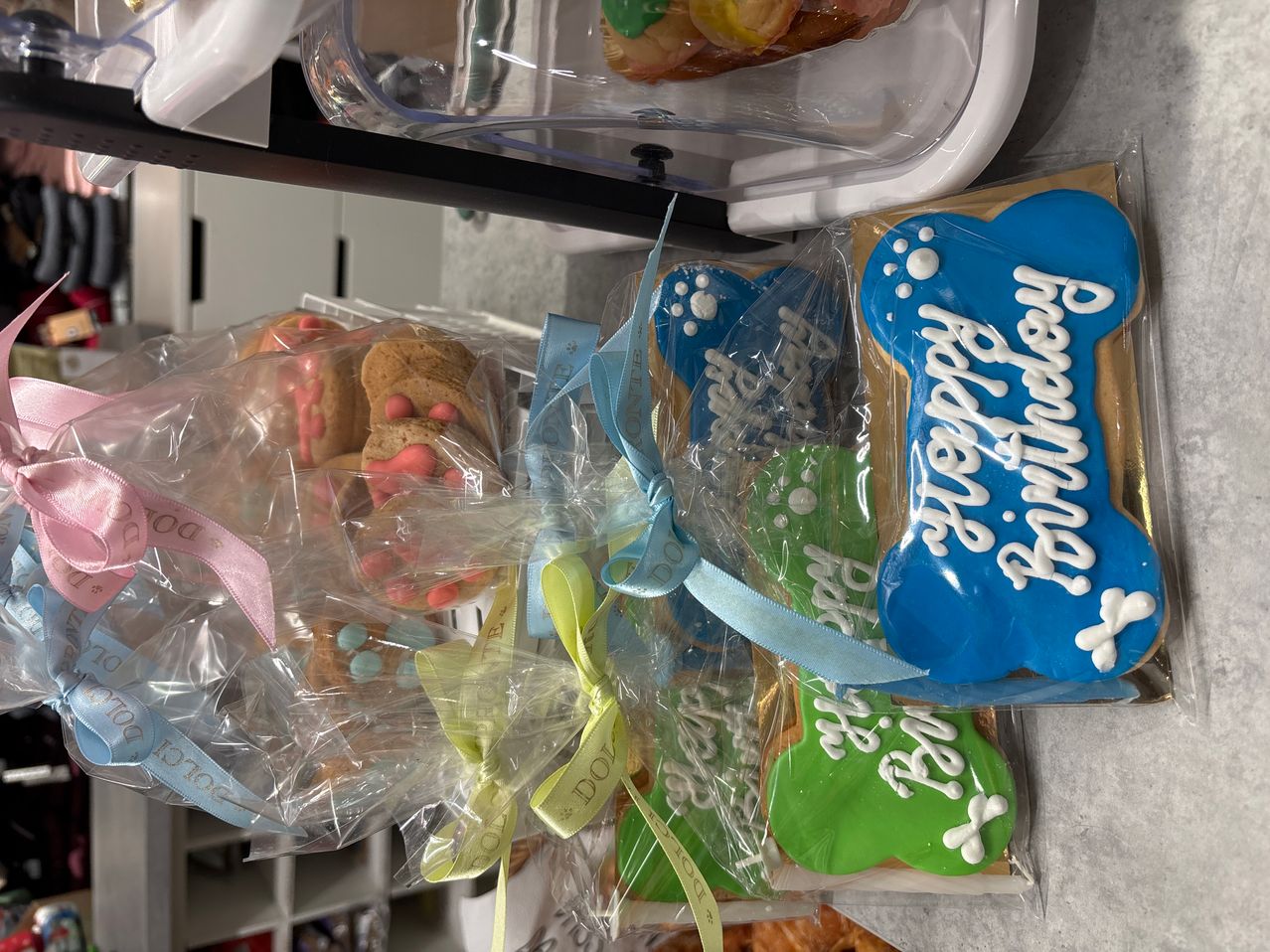 Mehrere Tüten mit Keksen stehen auf einem Tresen. Jede Tüte hat ein Band. Eine Tüte sagt Happy Birthday Rex. Eine andere sagt Happy Birthday Rocco. Kekse sind in Plastik eingewickelt.