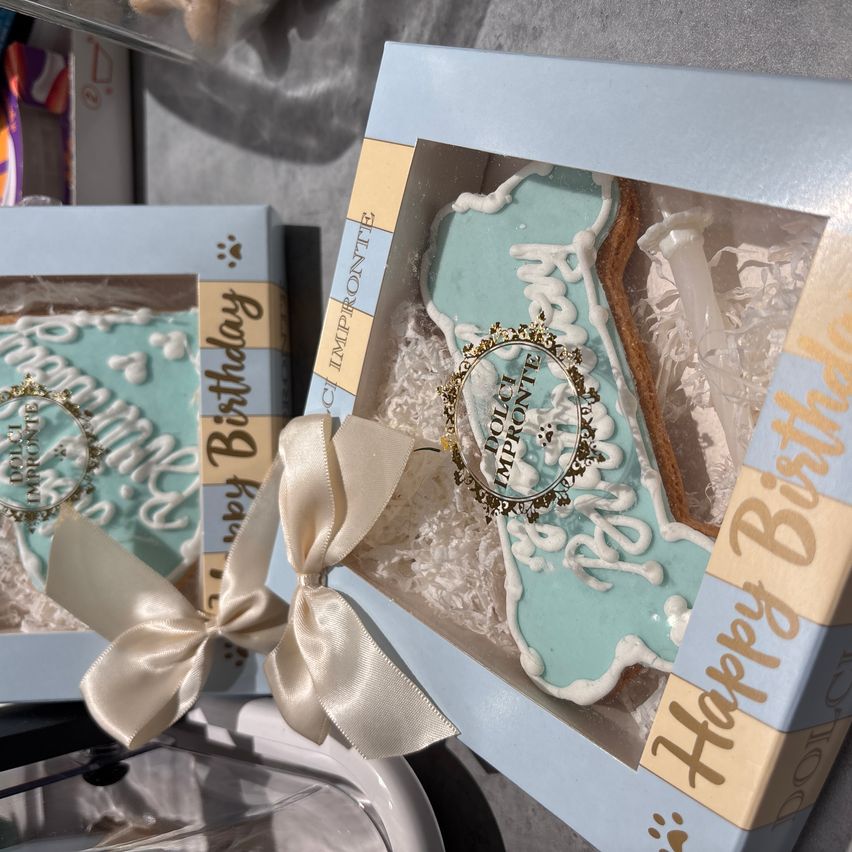 Zwei blaue und weiße Hundegeburtstagskuchen sind in Schachteln. Einer hat ein Band und die Aufschrift 'Happy Birthday'. Der andere hat ein goldenes Etikett mit dem Text 'Imp'. Beide sind in blauen Schachteln mit Bändern.