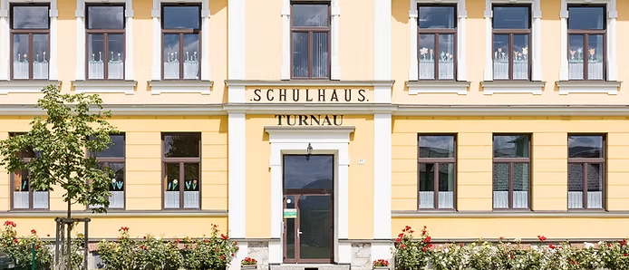 Ein gelbes Gebäude mit der Aufschrift Schulhaus Turnau oben. Es hat viele Fenster und eine große Eingangstür. Das Gebäude ist von einem gepflegten Garten mit roten Blumen umgeben.