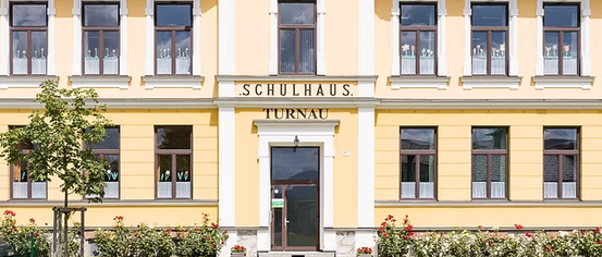 Ein gelbes Gebäude mit der Aufschrift Schulhaus Turnau oben. Es hat viele Fenster und eine große Eingangstür. Das Gebäude ist von einem gepflegten Garten mit roten Blumen umgeben.