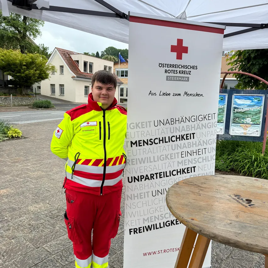Ein Mann in einem gelben und roten Uniform steht unter einem Baldachin mit einem Schild für das Österreichische Rote Kreuz.