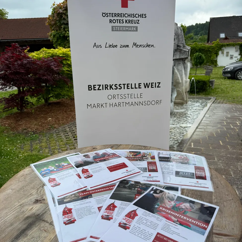 Ein Rotes-Kreuz-Schild steht im Garten und wirbt für die 'Bezirksstelle Weiz' in Hartmannsdorf. Broschüren und Flyer liegen auf einem Tisch davor.