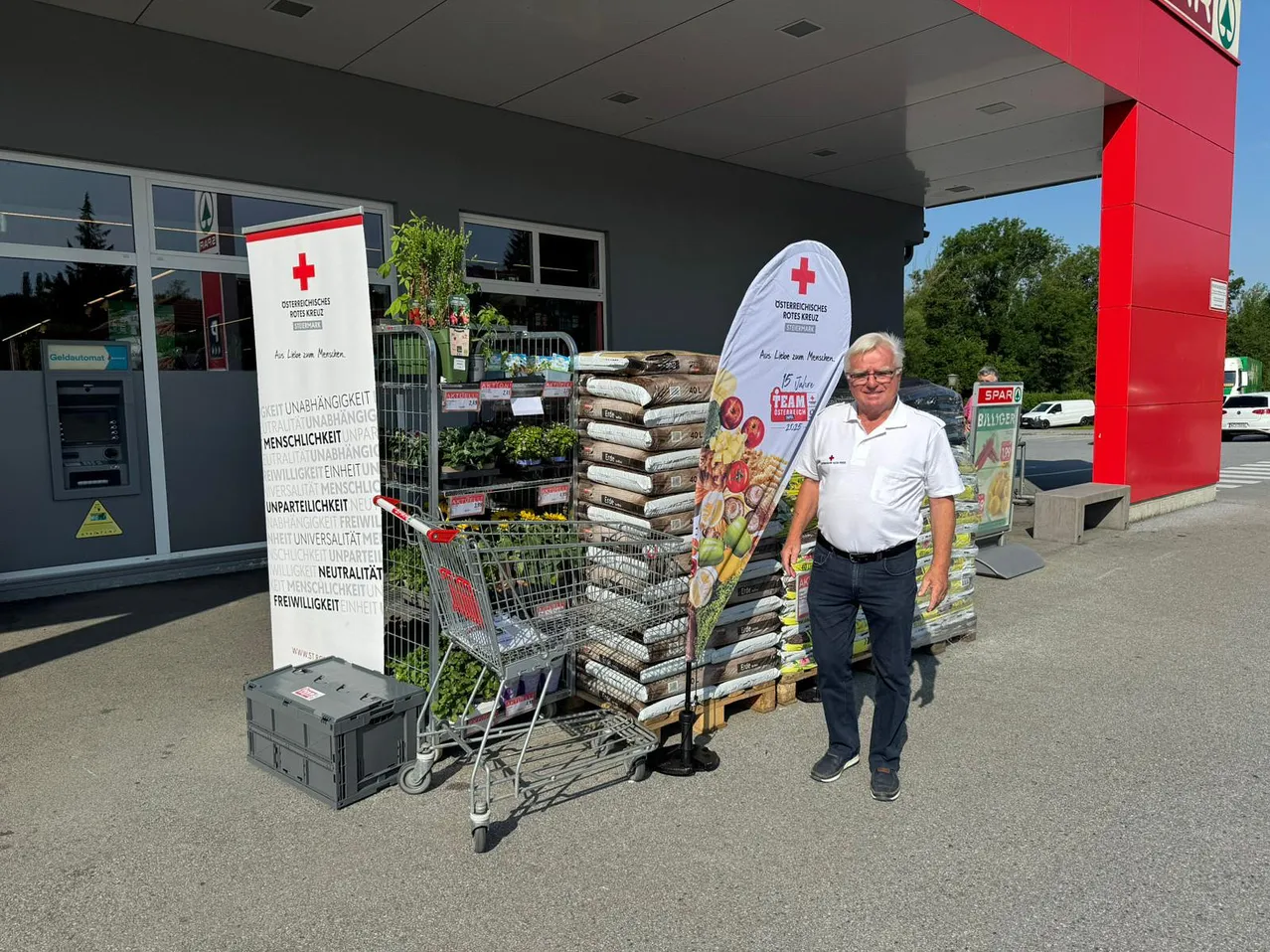 Ein Mann steht neben einem Einkaufswagen und Bannern, mit gestapelten Bodensäcken und einem roten Kreuzbanner hinter ihm.