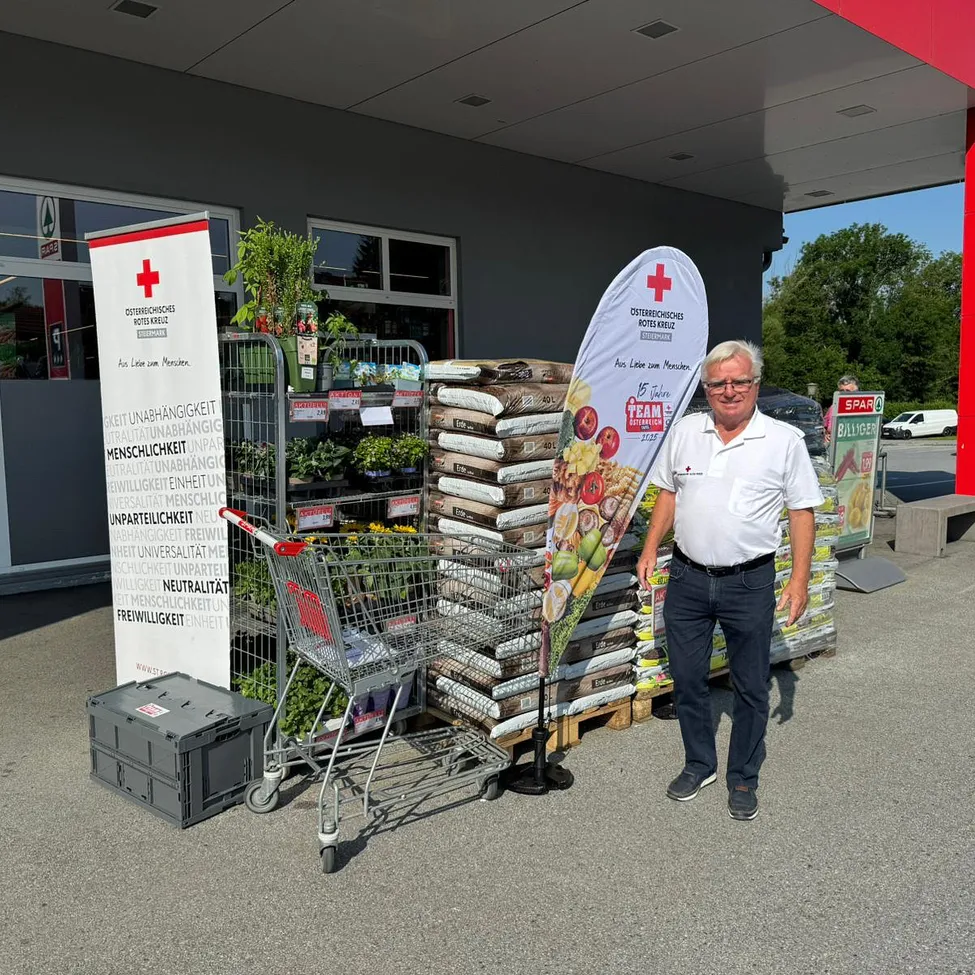 Ein Mann steht neben einem Einkaufswagen und Bannern, mit gestapelten Bodensäcken und einem roten Kreuzbanner hinter ihm.