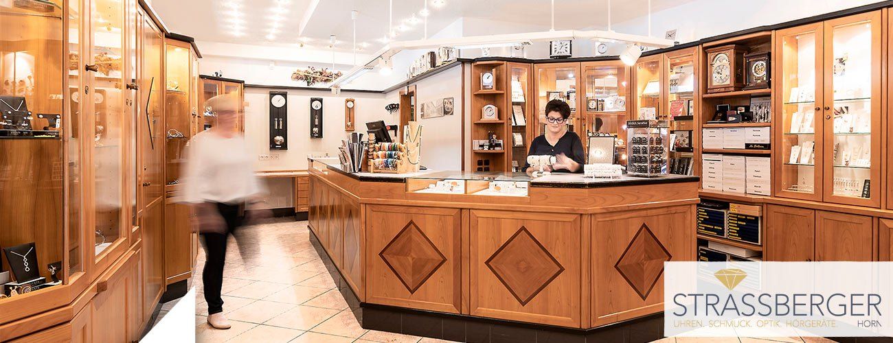 Eine Frau mit Brille steht hinter einem hölzernen Empfangstresen in einem Juwelierladen. Der Laden hat Holzschränke, Uhren und Regale. Eine Person in Weiß geht im Hintergrund.
