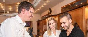 Ein Mann in einem weißen Hemd hält die Hände eines Mannes in einem schwarzen T-Shirt, während eine Frau in einem weißen Blusenhemd hinter ihnen steht. Sie alle lächeln.