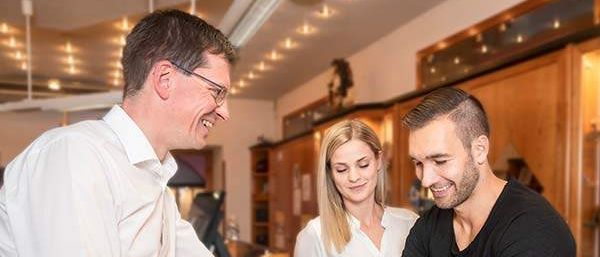 Ein Mann in einem weißen Hemd hält die Hände eines Mannes in einem schwarzen T-Shirt, während eine Frau in einem weißen Blusenhemd hinter ihnen steht. Sie alle lächeln.