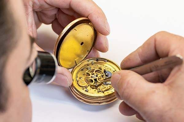 Eine Person benutzt eine Lupe, um eine goldene Taschenuhr zu inspizieren und zu reparieren, die sie in den Händen hält.
